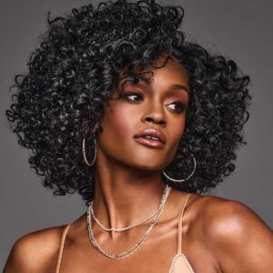 Portrait of a woman with a curly synthetic wig, wearing hoop earrings, a necklace, and a beige top, against a gray background.