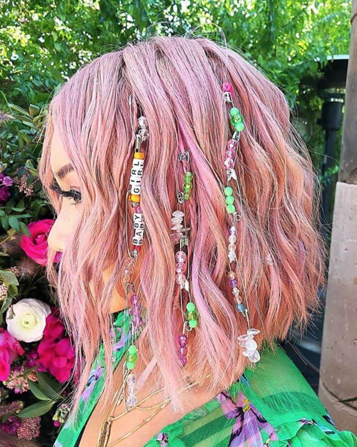 A person with short pink hair adorned with colorful beads and charms stands outside near vibrant plants and flowers.