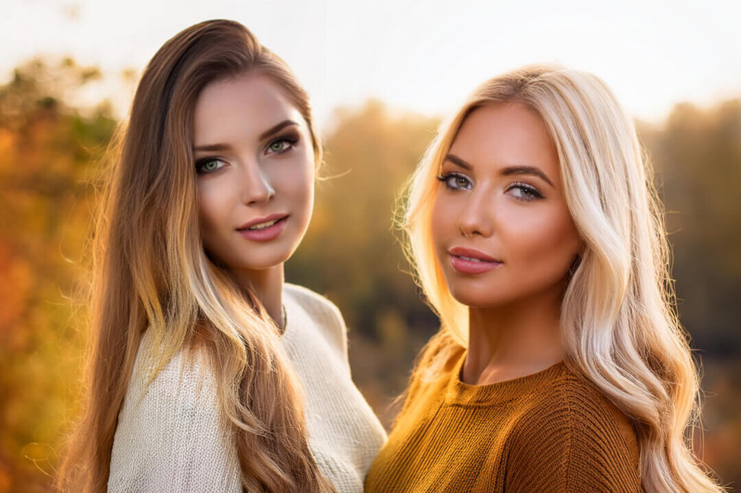 Two women with long hair, one brunette and one blonde, stand outdoors in fall, dressed in sweaters.